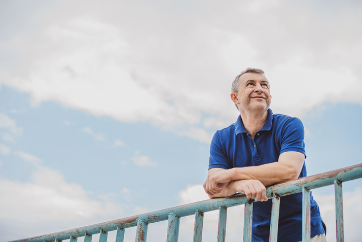 Lifelong learning po 50. roku życia - zdjęcie uśmiechniętego mężczyzny 50+ opartego o barierkę, nad nim zachmurzone niebo.