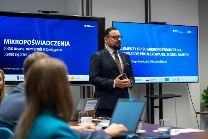 Grupa Doradcza ds. Mikropoświadczeń - spotkanie. Michał Nowakowski przemawia, w tle ekrany z nazwą projektu, z przodu uczestnicy spotkania